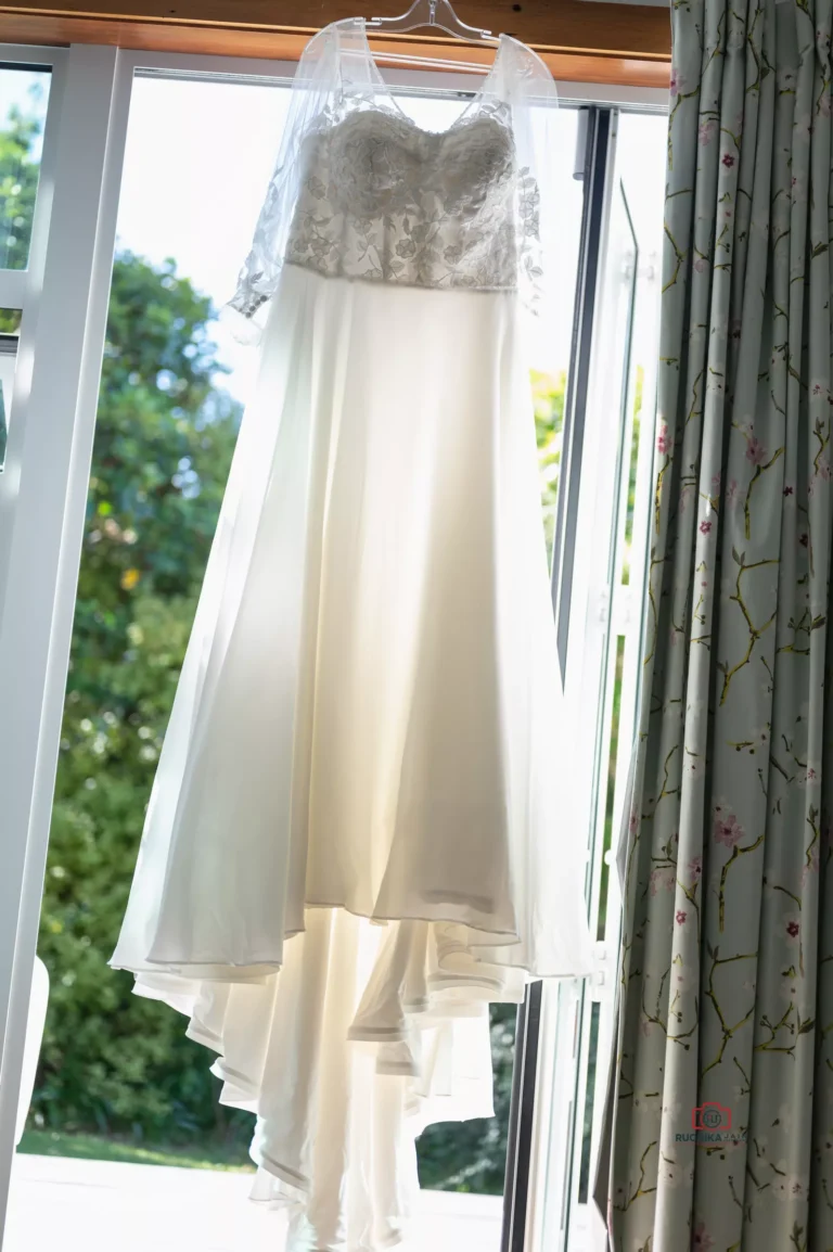 A stunning bridal gown with delicate lace details and a flowing skirt, hanging by a sunlit window with floral-patterned curtains