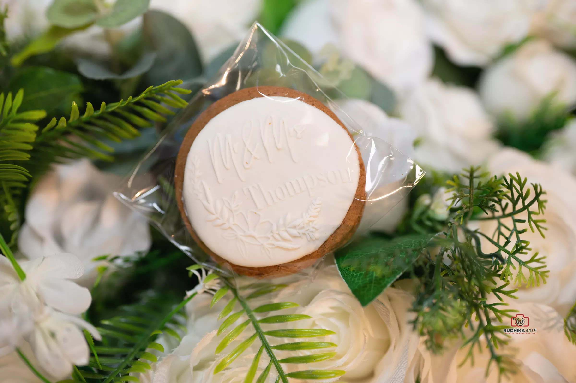 Wedding favor cookie with a floral embossed design, personalized text, and a backdrop of white roses and green foliage.