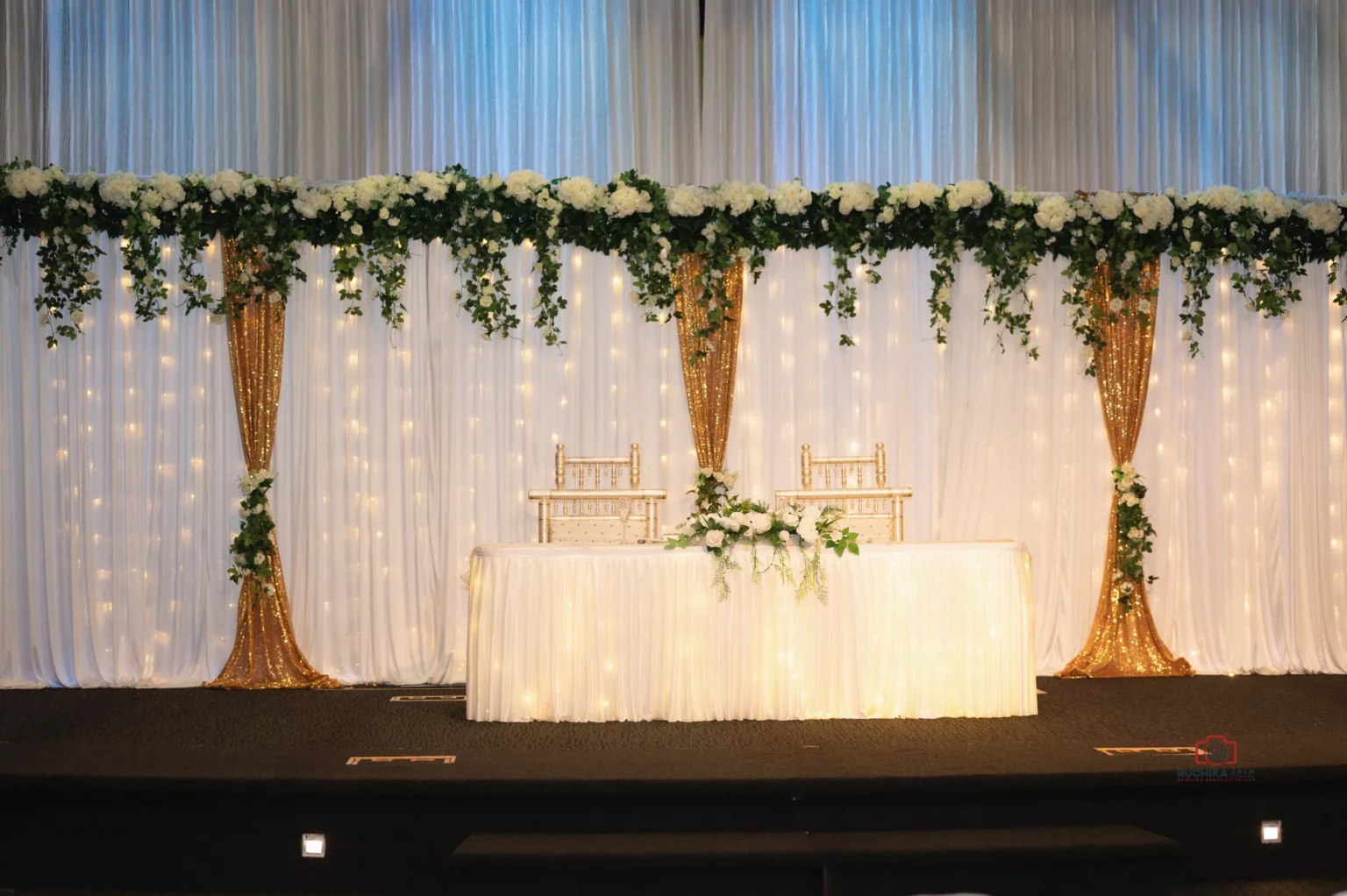 Wedding reception stage setup with white drapes, gold accents, floral garlands, and two chairs behind a decorated table