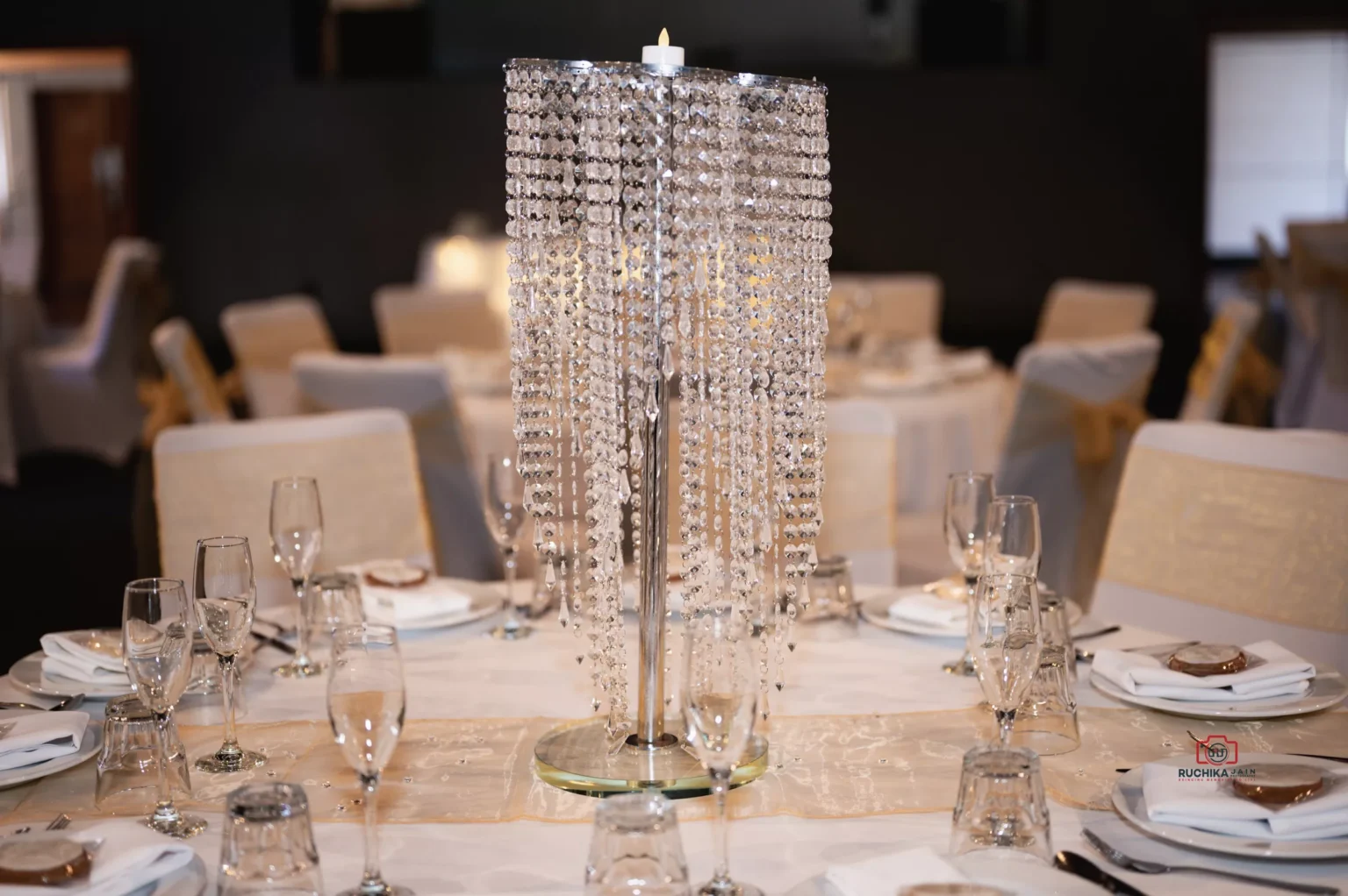 A beautifully decorated wedding reception table featuring a luxurious crystal centerpiece with cascading details, surrounded by white tableware, glasses, and gold-accented chair covers.