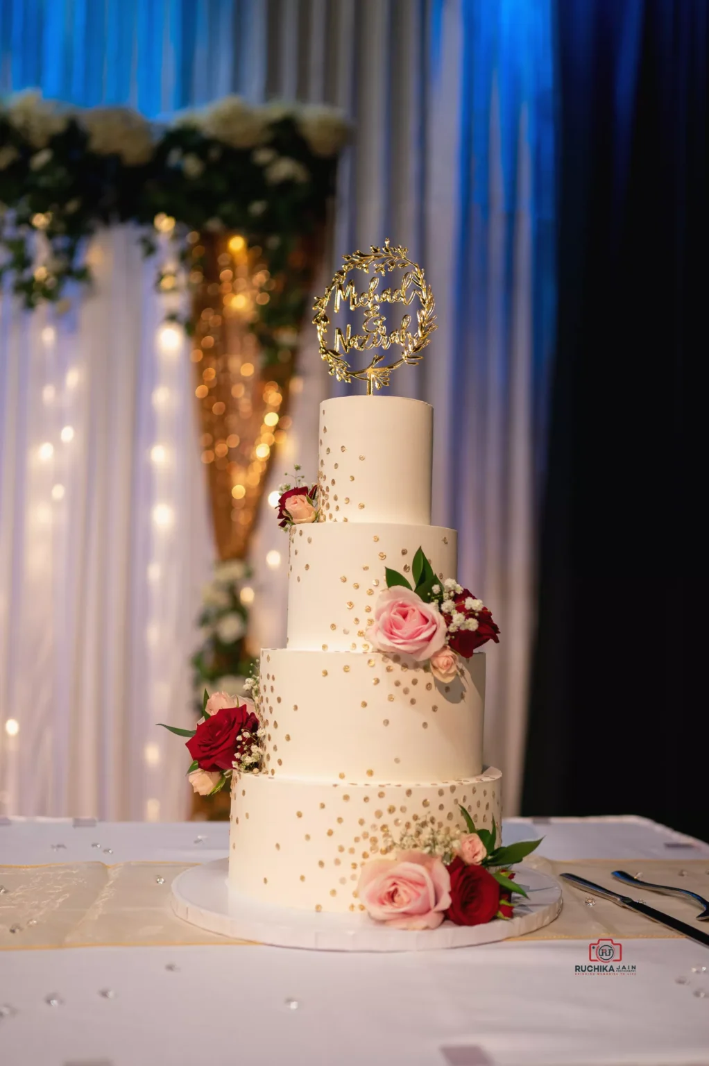 Three-tiered white wedding cake adorned with red and pink roses and a custom topper reading "Mr & Mrs"