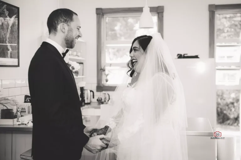 Black-and-white image capturing an emotional first look moment between a bride in a veil and a groom in a tuxedo, sharing smiles and holding hands