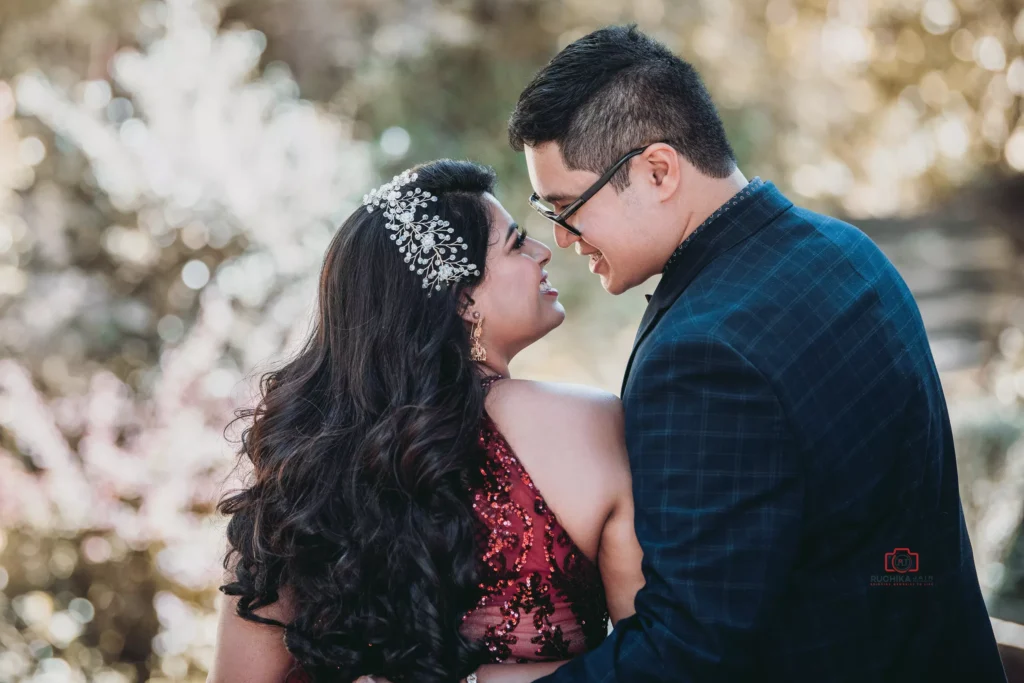 Engaged couple embracing outdoors, gazing into each other's eyes with smiles, with the woman wearing a jeweled hairpiece and red dress