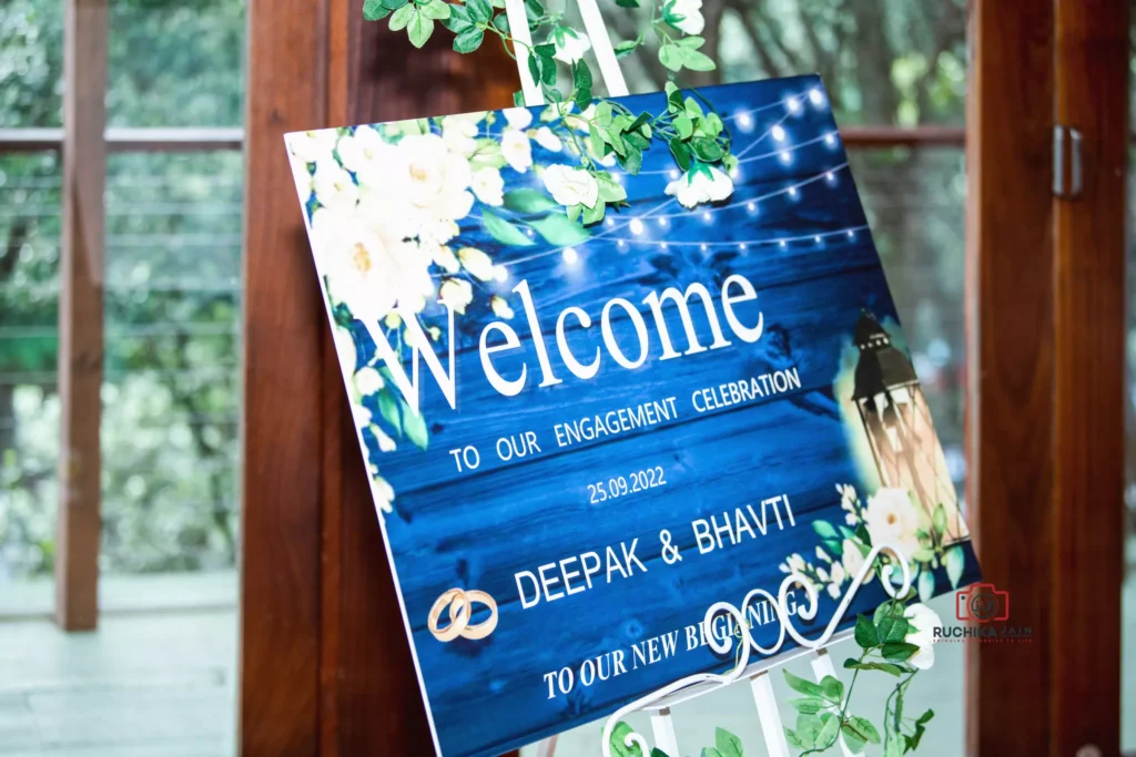 A blue-themed welcome sign for Deepak and Bhavti's engagement celebration, decorated with white flowers and lantern design