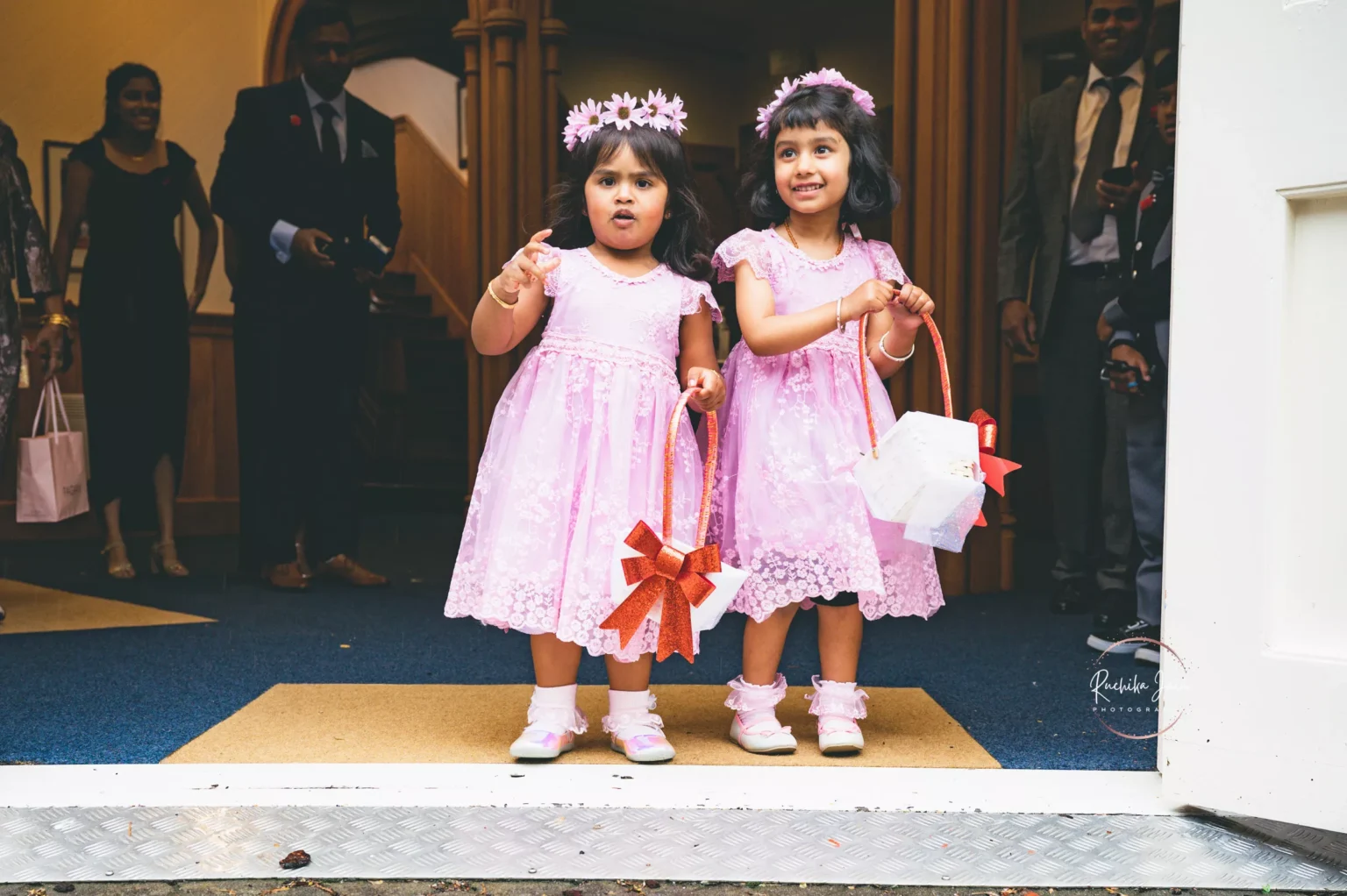 Two young flower girls in matching pink dresses and flower crowns hold baskets decorated with ribbons, standing at the church entrance