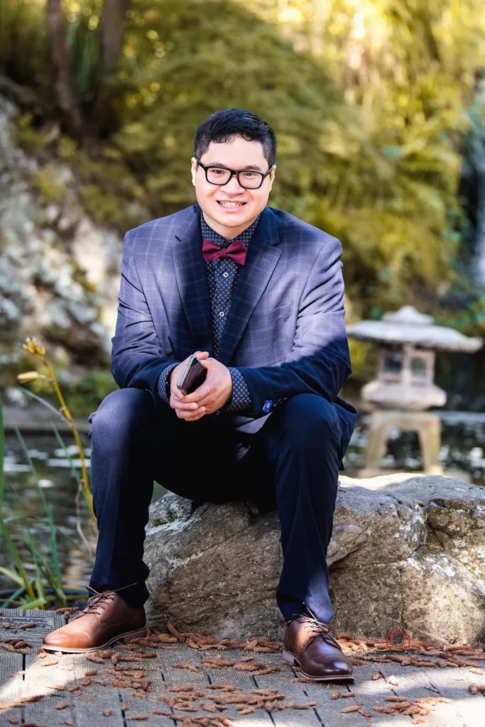 Groom in a navy suit with a red bow tie, sitting on a rock in a garden setting, smiling towards the camera