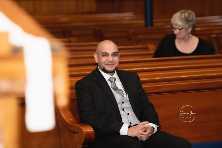 Groom in a black suit and grey vest seated in a church pew, smiling warmly