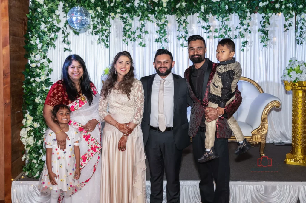 A group photo featuring a joyous gathering at a beautifully decorated event. The image showcases family and friends posing against a lush green and white floral backdrop, with a golden and white throne-style chair as part of the setting.