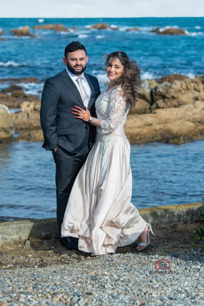 Bride and groom standing together by the ocean in Wellington, New Zealand, with rocky coastline and blue water in the background