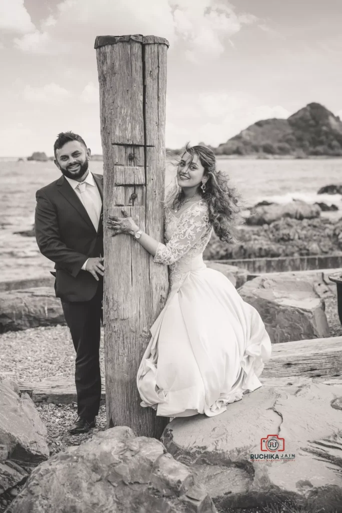 Bride and groom posing by a weathered wooden post on a rocky Wellington beach, with the ocean and hills in the background