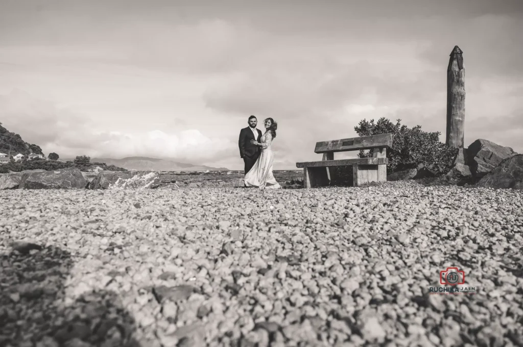 A black-and-white photograph of a couple embracing during their pre-wedding shoot on a rocky shoreline with a bench and scenic views in the background.