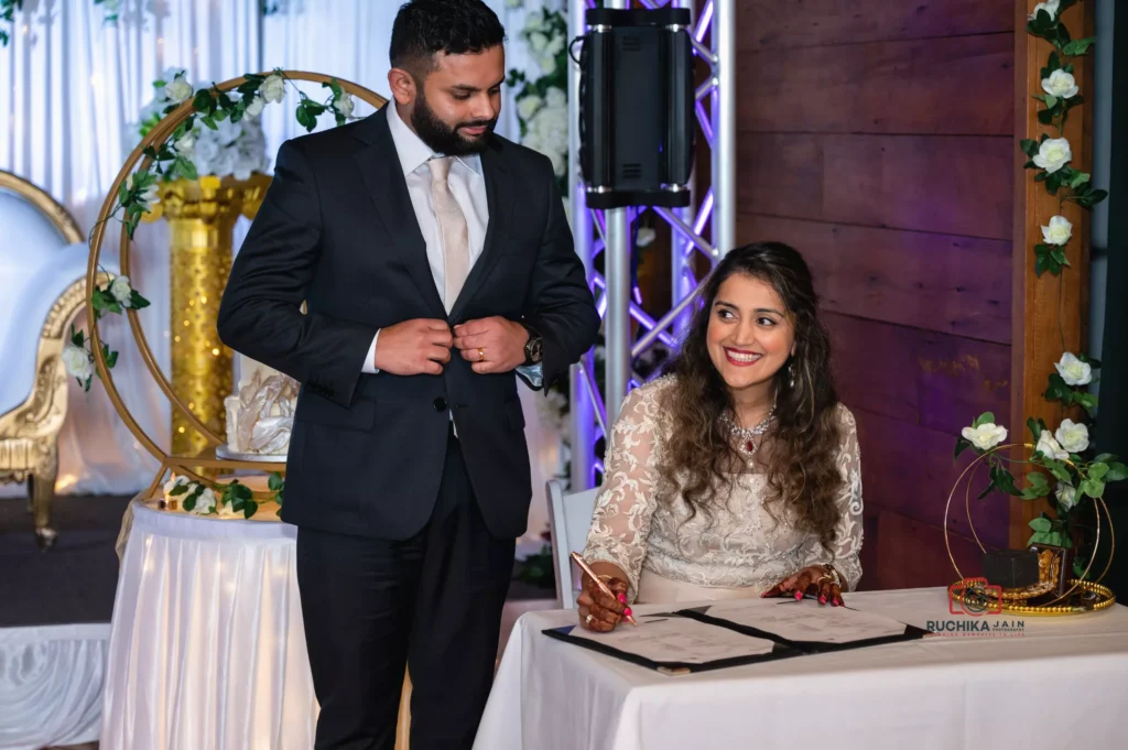 A bride signs the marriage certificate with a joyful smile while the groom looks on lovingly. The scene is set with elegant gold and floral decor, representing the formal yet intimate moment of their union.