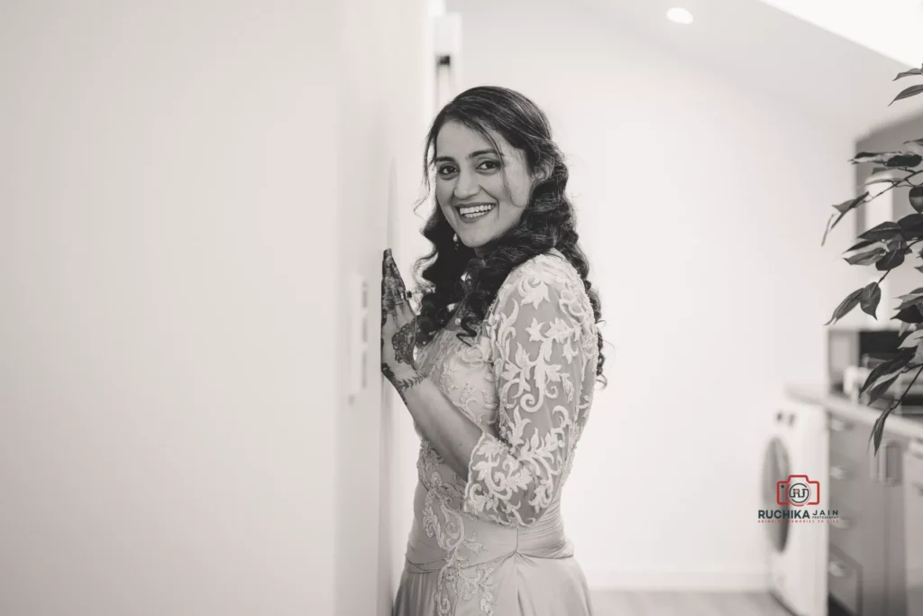 Bride smiling in a lace gown with intricate patterns, standing indoors and looking toward the camera