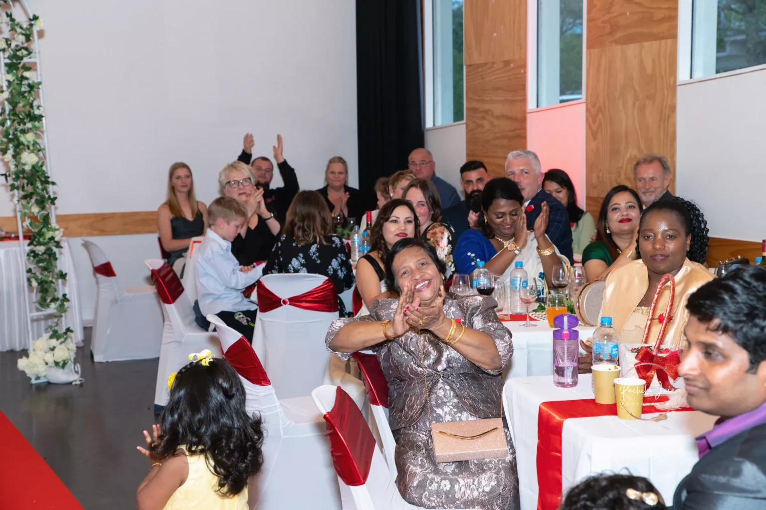 Guests at a wedding reception applauding and smiling, seated at decorated tables with red and white accents