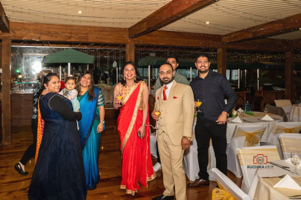 Group of wedding guests, including women in traditional attire, posing with drinks at a warmly lit reception venue