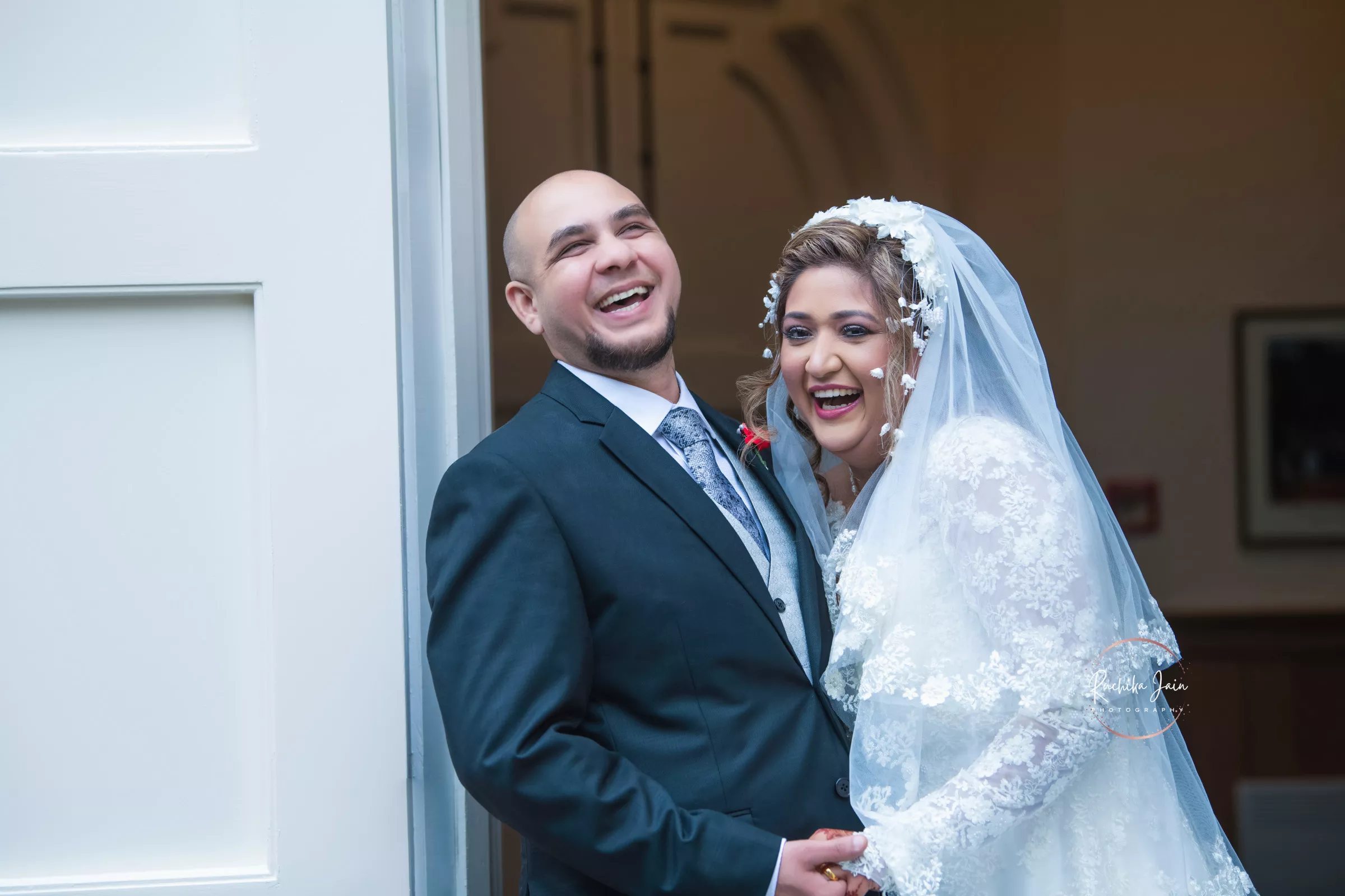 A happy bride in a lace wedding gown with a veil and her groom in a dark suit share a laugh, radiating joy and love on their wedding day.