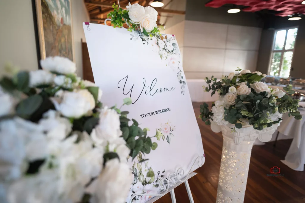 A beautifully designed wedding welcome sign adorned with floral decorations, surrounded by white and green floral arrangements, captured at Silver Stream Retreat, Upper Hutt