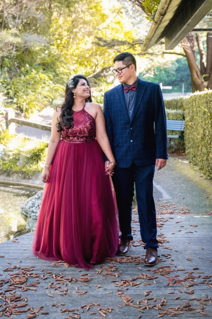 Zara, wearing an elegant burgundy gown with intricate lace details, walking hand in hand with Andrew, dressed in a navy blue checkered suit with a red bow tie, sharing a joyful moment in a lush garden setting.