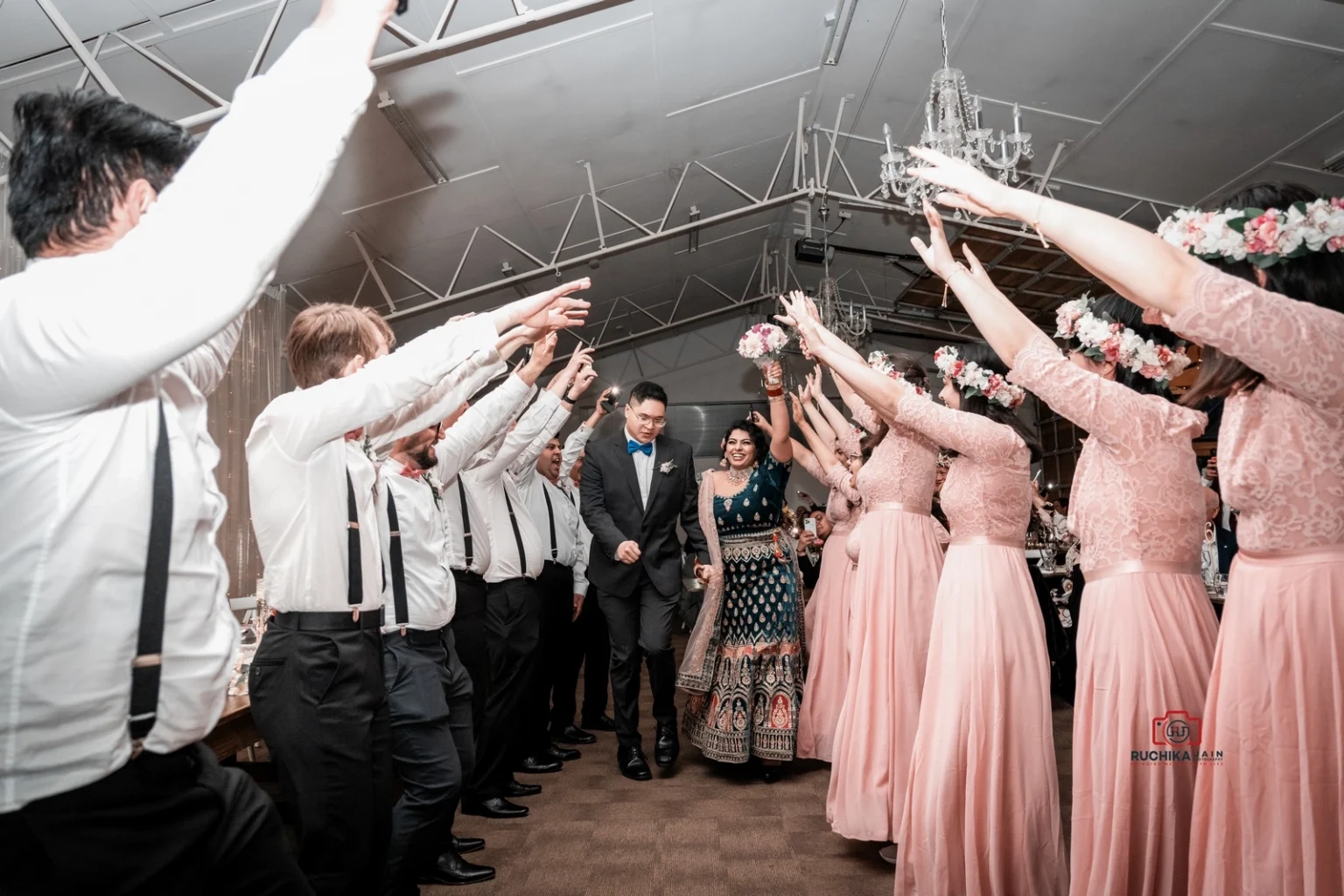 Bride and groom entering the reception through a tunnel of raised arms formed by bridesmaids in pink dresses and groomsmen in suspenders