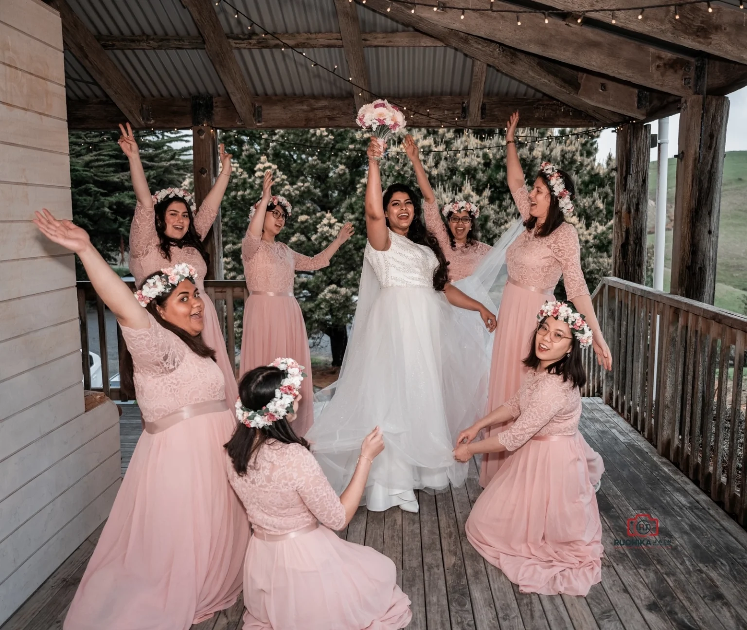 Bride in a white dress holding her bouquet high with bridesmaids in pink dresses and floral crowns joyfully raising their arms on a rustic porch