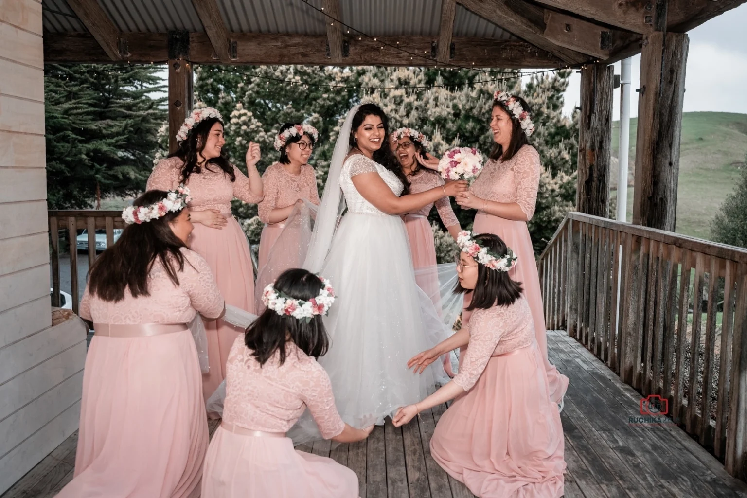 Bride holding her bouquet and laughing with her bridesmaids in pink dresses and floral crowns on a wooden porch, surrounded by greenery