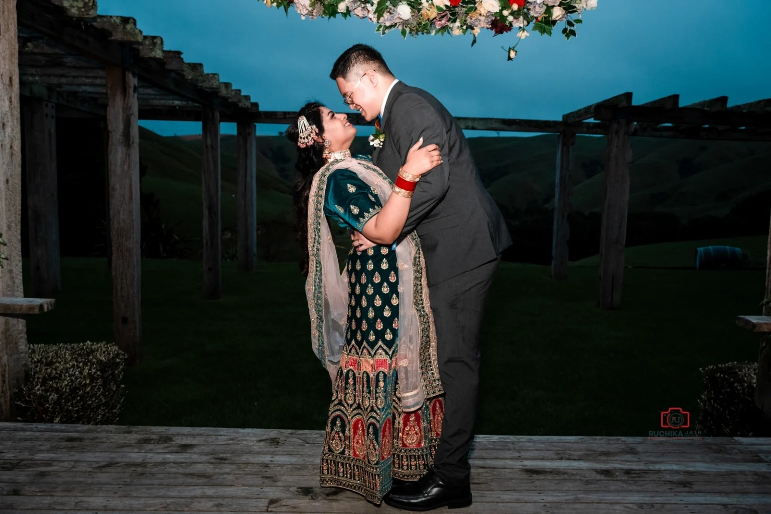 Bride in traditional green and gold attire and groom in a suit share an embrace under a floral arch outdoors