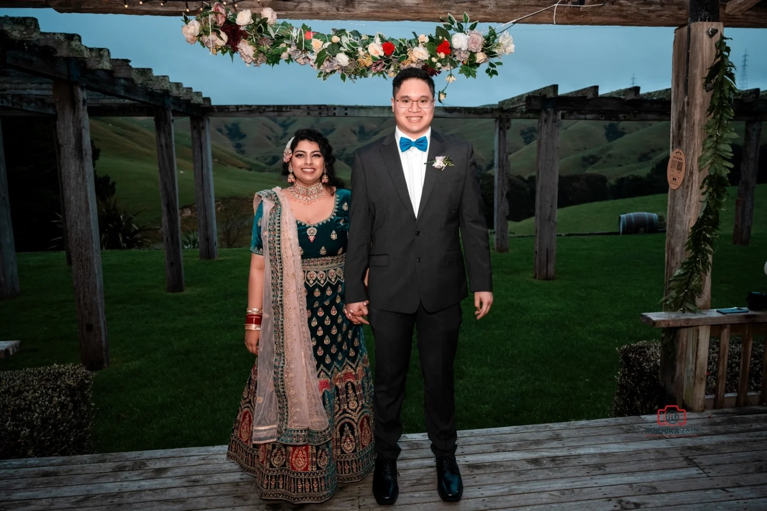 Bride in a traditional green and gold outfit with groom in a dark suit, standing under a floral arch with a scenic outdoor background