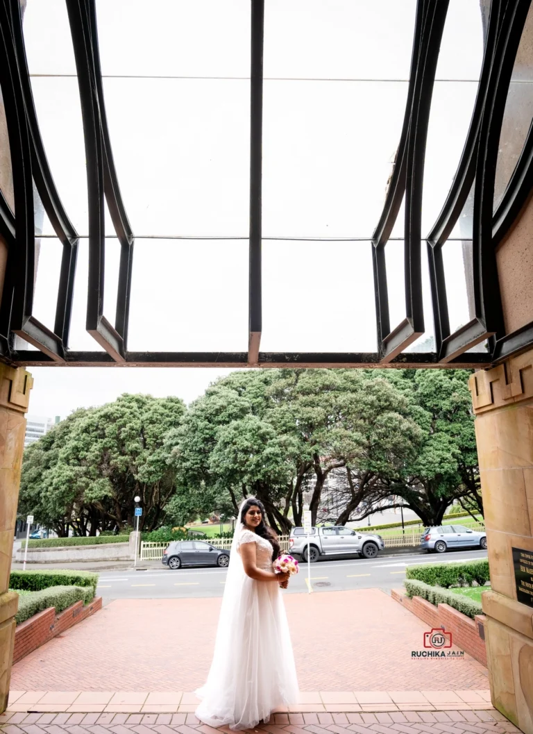 Bride in white dress holding a bouquet, standing under a large architectural archway with trees and street in the background