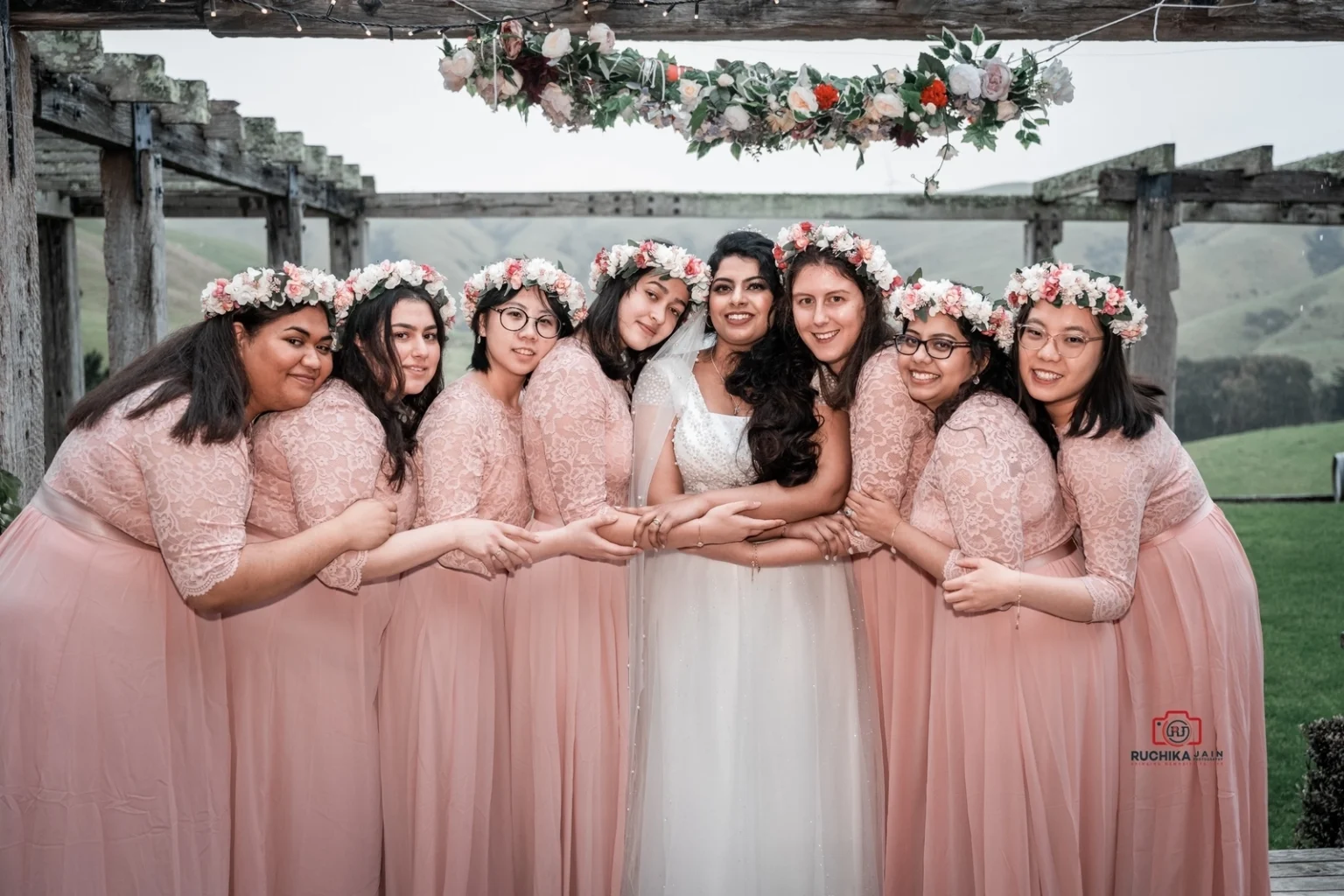 Bride in a white dress surrounded by eight bridesmaids in pink dresses and floral crowns, standing closely together under a pergola with floral decorations