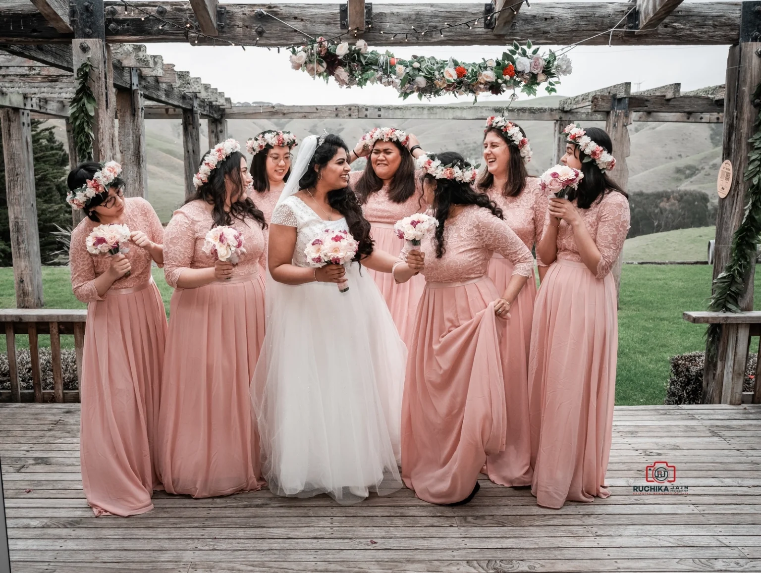 Bride standing with her bridesmaids dressed in matching pink gowns, laughing together under a decorated wooden pergola