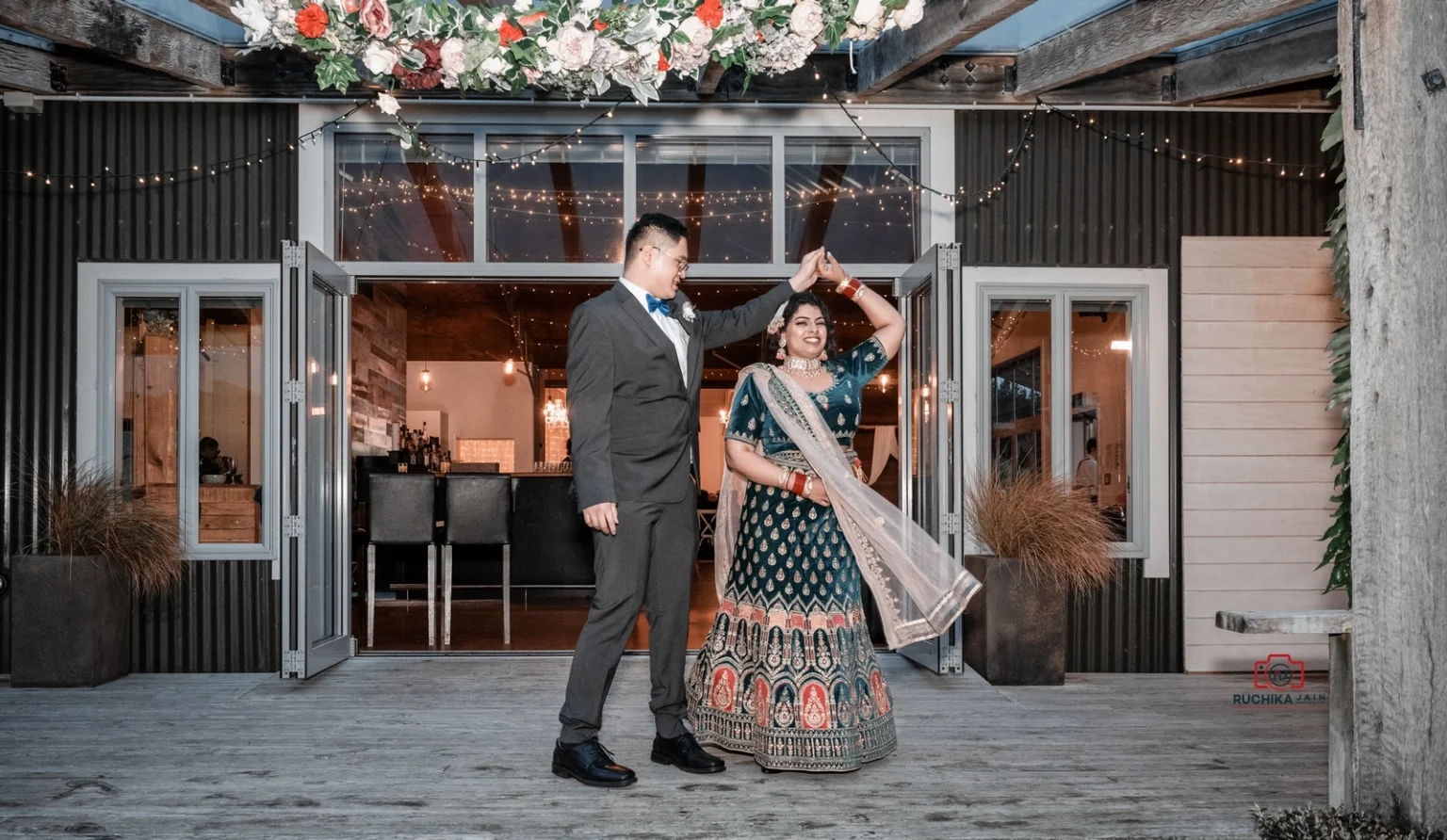 Groom twirls bride in traditional green and gold lehenga outside a venue with open doors and string lights