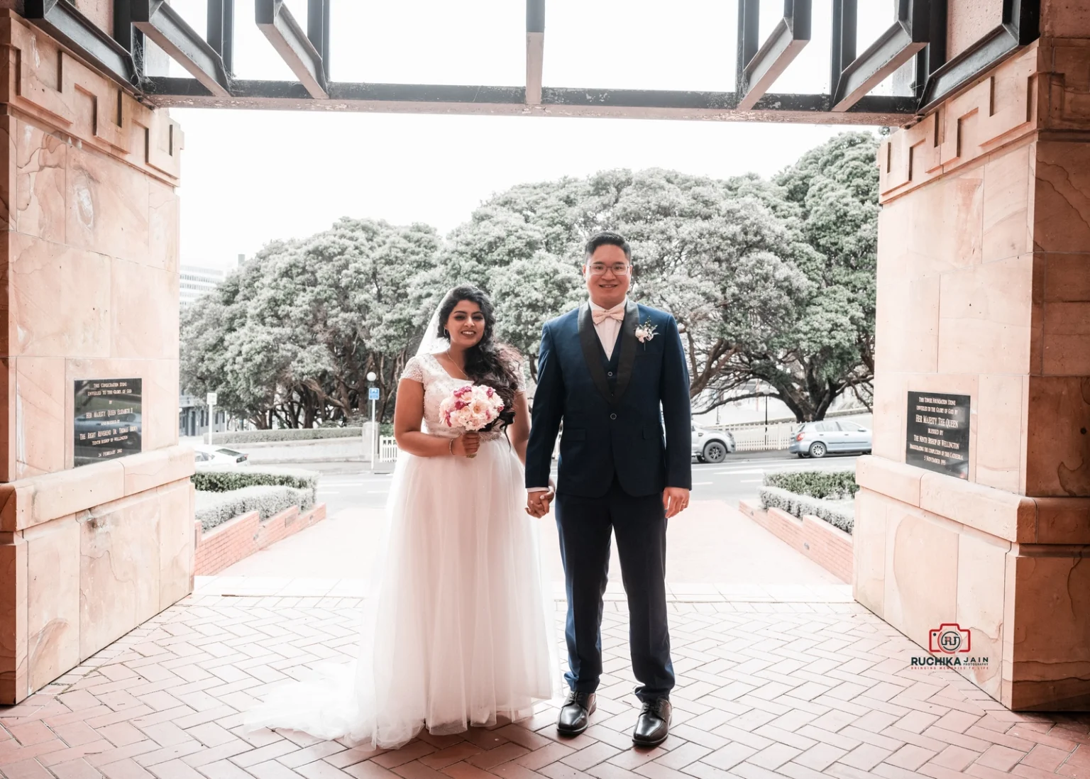 Bride in white dress holding a bouquet stands beside groom in blue suit at an outdoor entrance, with greenery and trees in the background