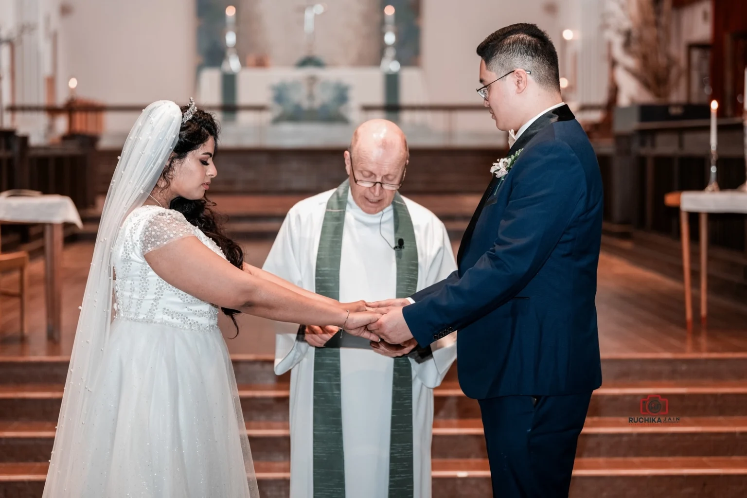 Bride and groom holding hands during their wedding ceremony in a church, with a priest standing between them, guiding the exchange of vows