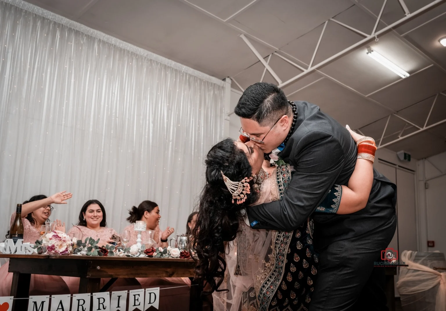 The groom leans in and kisses the bride during a romantic dance, with the bridesmaids smiling in the background.