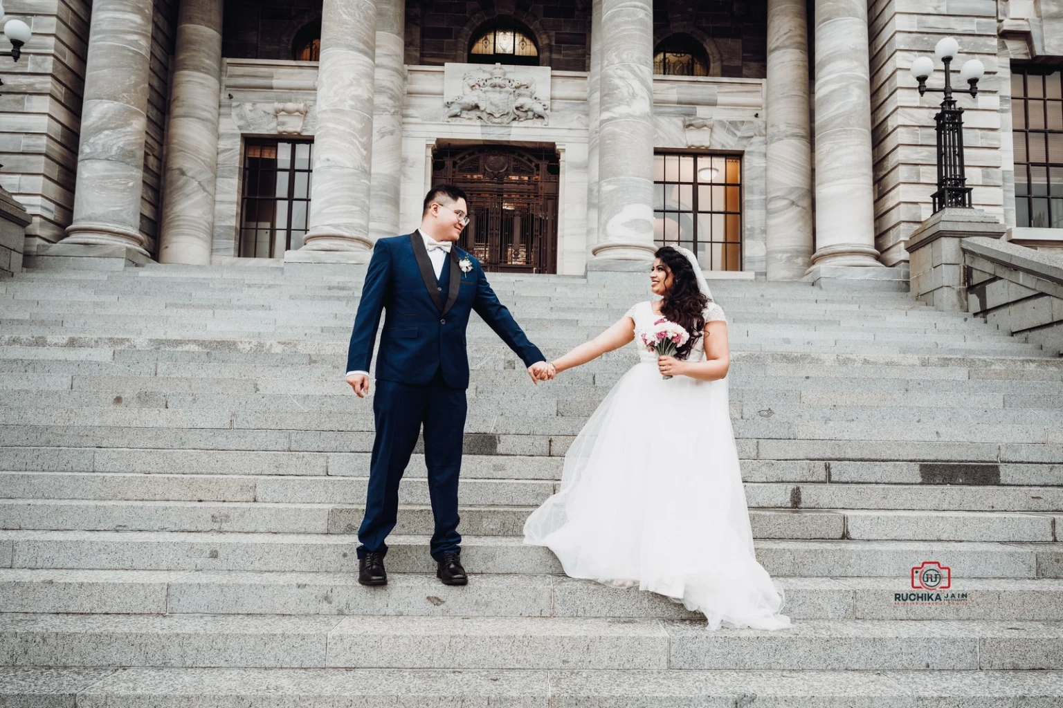 Bride and groom holding hands on the steps of a grand building with stone columns, smiling at each other