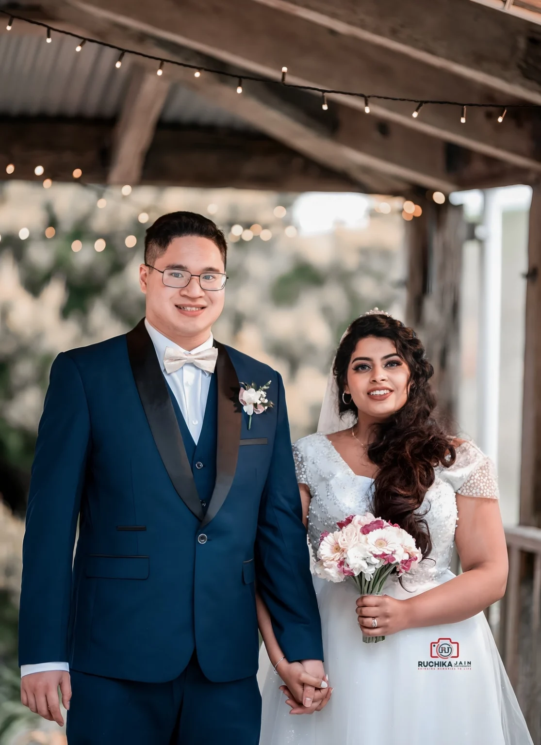 Bride and groom standing together, smiling and holding hands under string lights