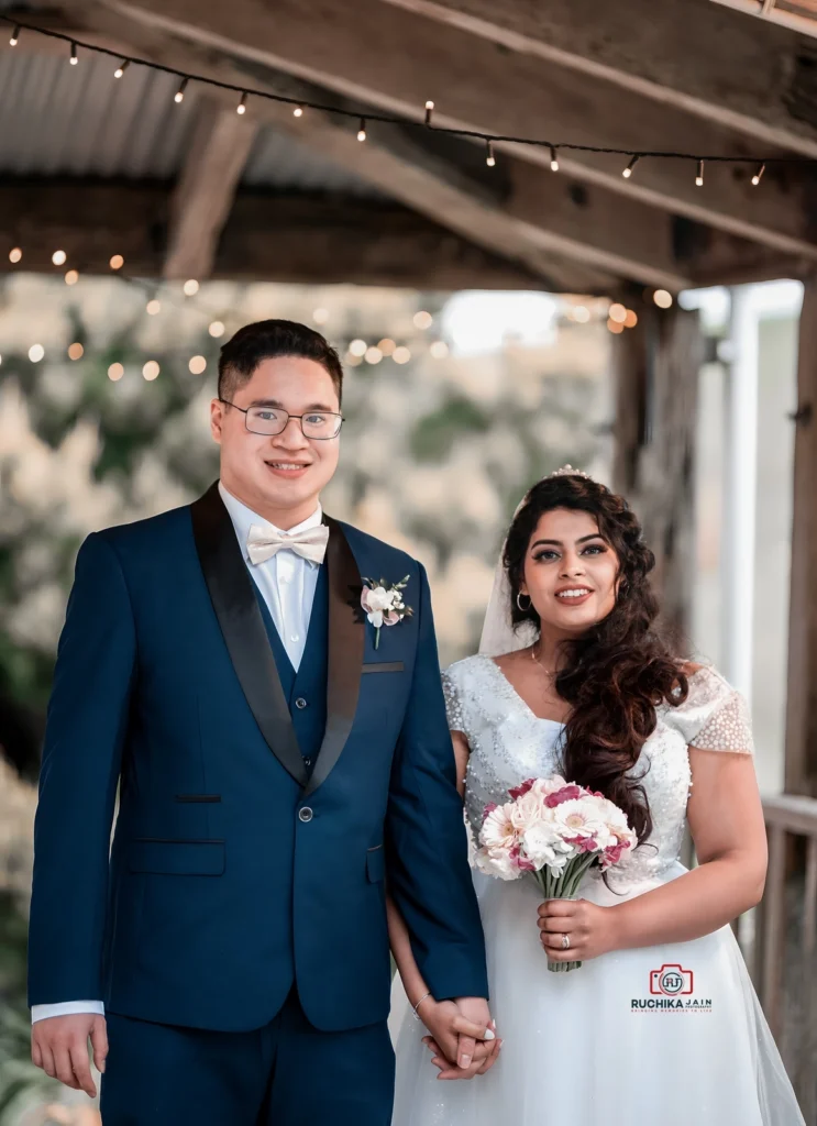 Bride and groom standing side by side holding hands, groom in a blue suit and bride in a white dress with a pink bouquet, under string lights