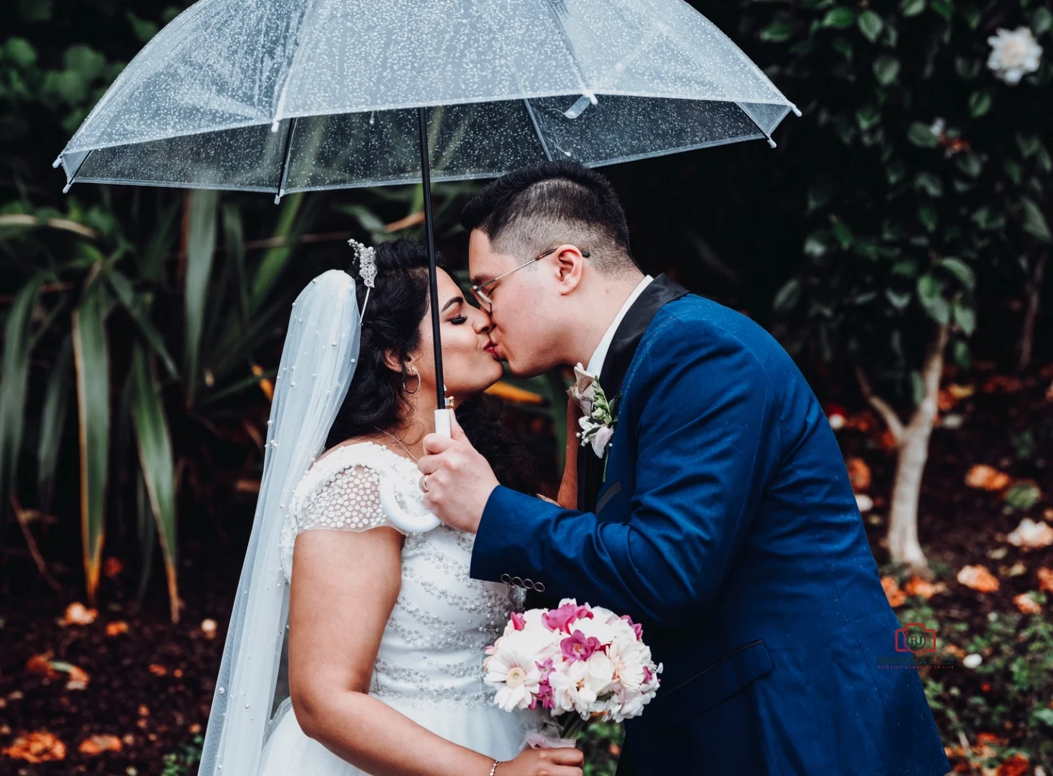 Bride and groom kissing under a clear umbrella on a rainy day, with the bride holding a bouquet of pink and white flowers