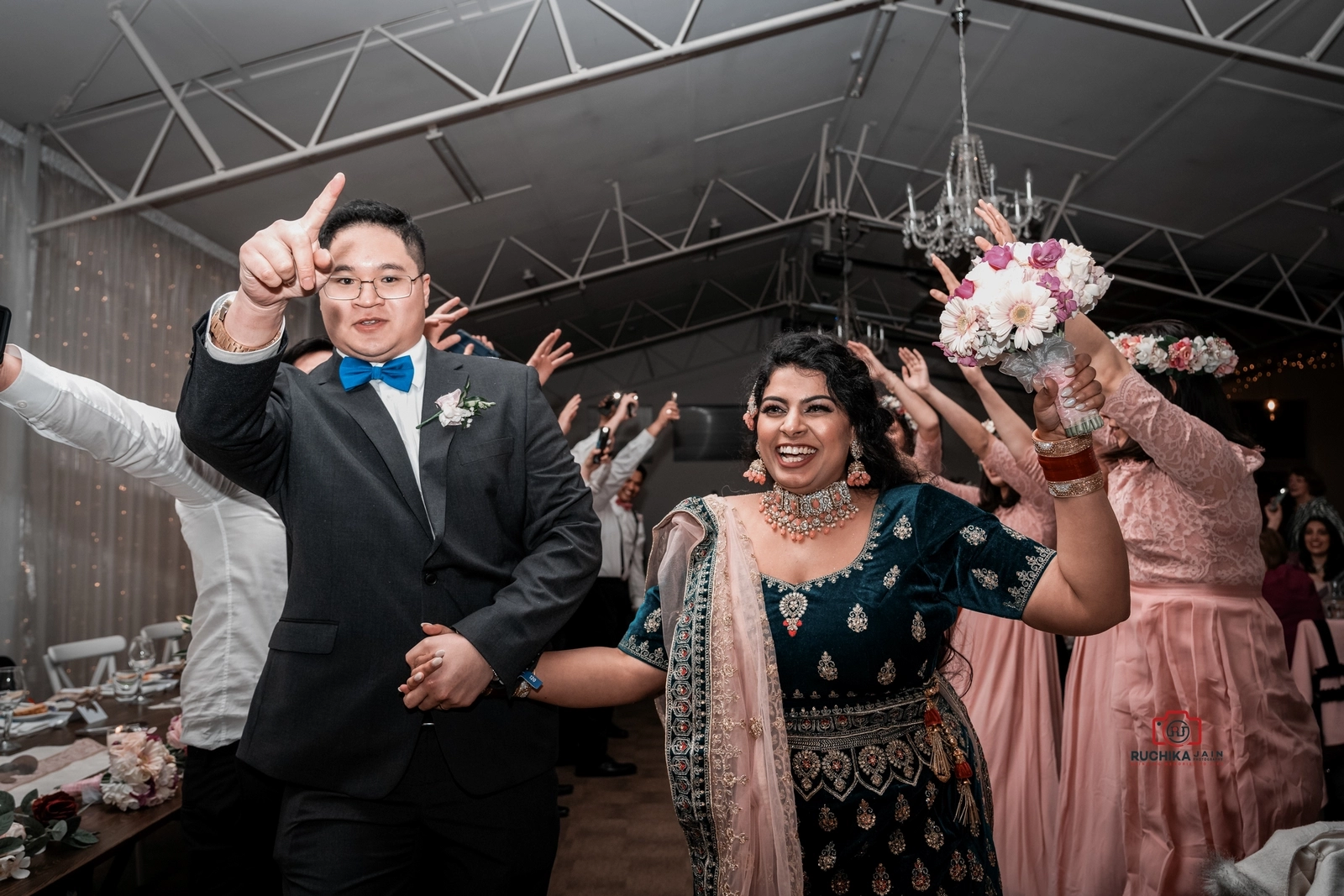 Smiling bride holding a bouquet and groom pointing, both walking into the reception as guests cheer in the background