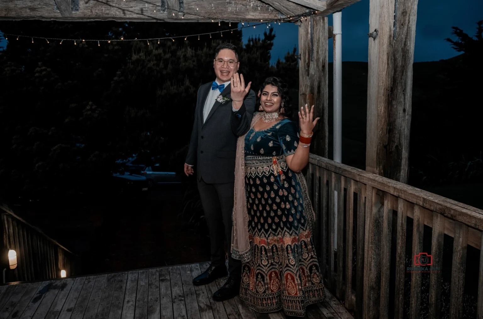 Bride and groom proudly display their wedding rings on an outdoor deck, with string lights overhead at dusk