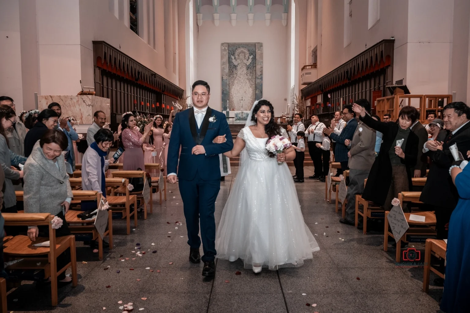 Bride and groom walk down the aisle together, smiling, as guests cheer and throw flower petals