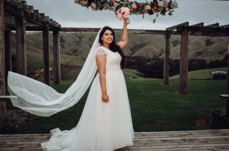 A bride in a white wedding dress holds her bouquet above her head, standing under a floral arch with rolling green hills in the background