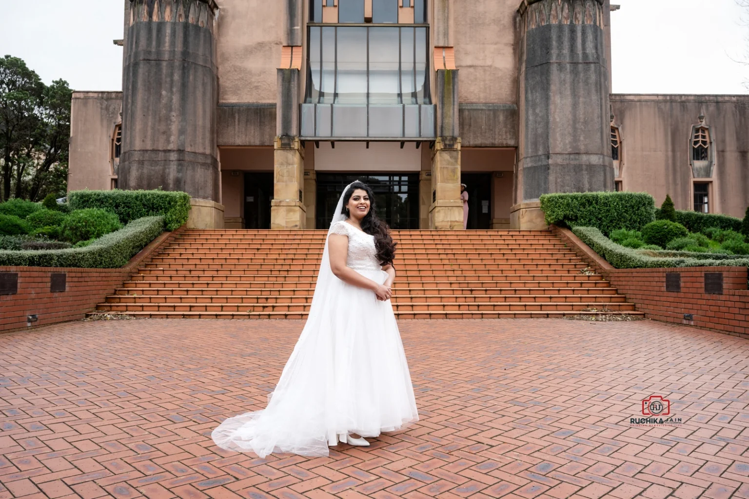 Bride in a flowing white dress standing in front of a grand building with large steps and manicured hedges