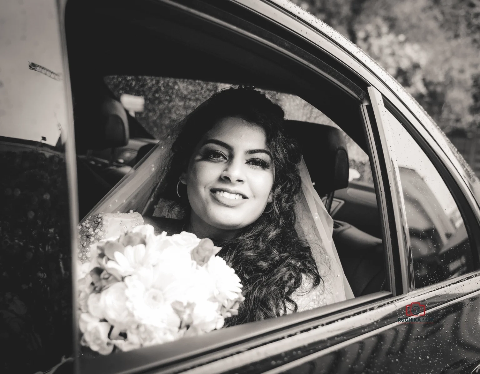 Bride smiling through the window of a car, holding a bouquet, with her veil visible in a black-and-white photograph