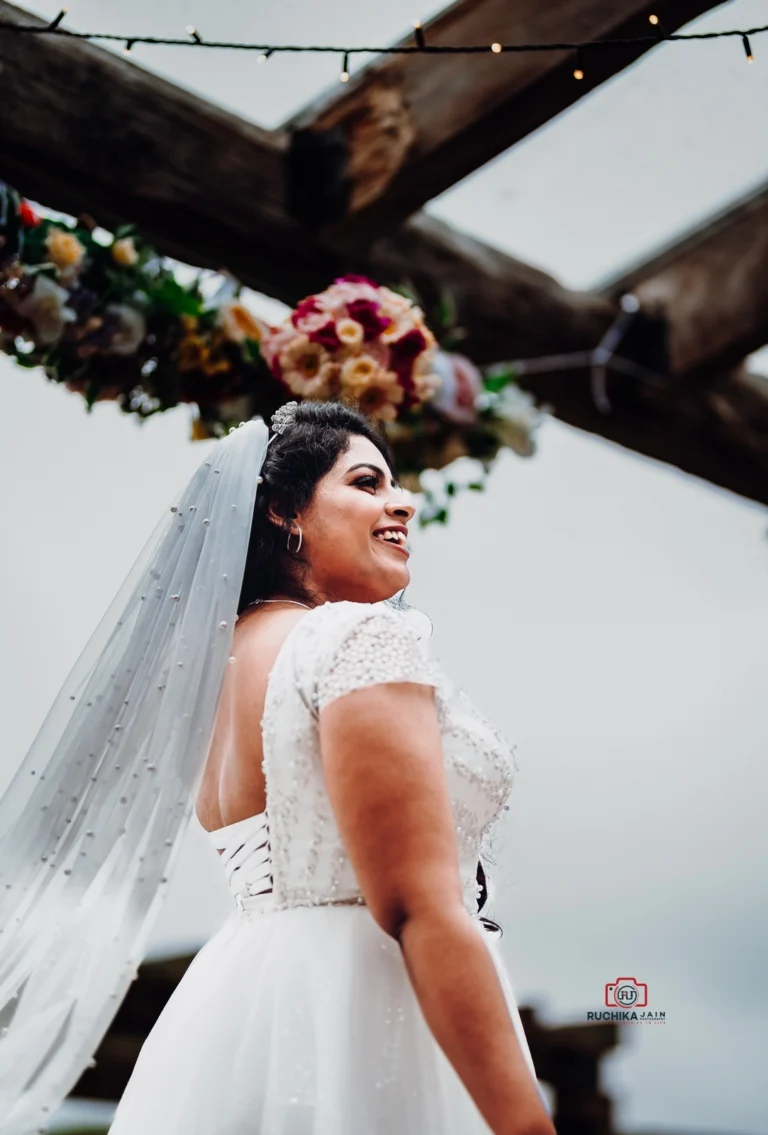 A close-up profile shot of a bride smiling under a floral arch. She wears a beaded white wedding dress with cap sleeves and a veil adorned with pearls