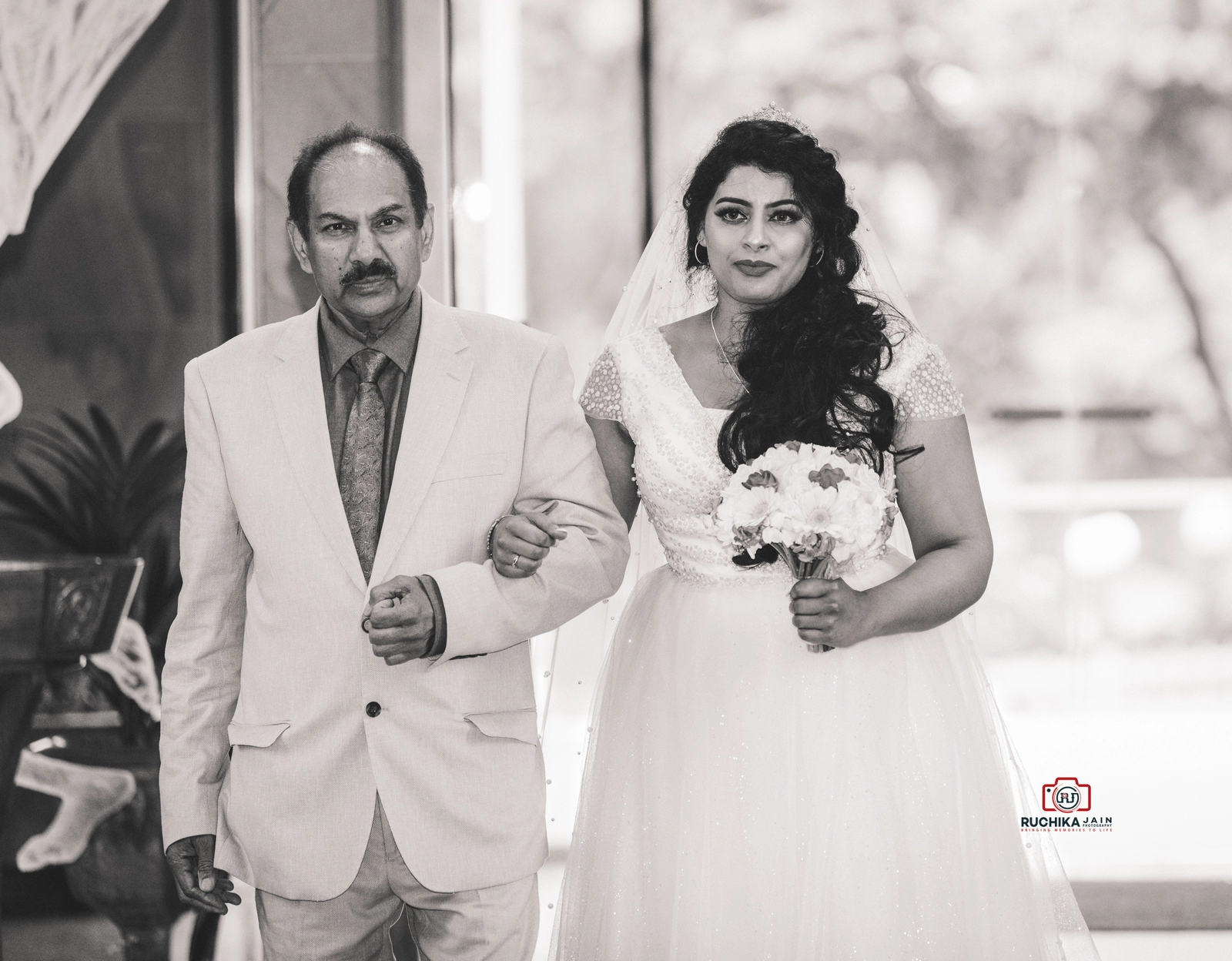 A black-and-white photo of a bride holding a bouquet, walking down the aisle arm-in-arm with her father at a wedding ceremony