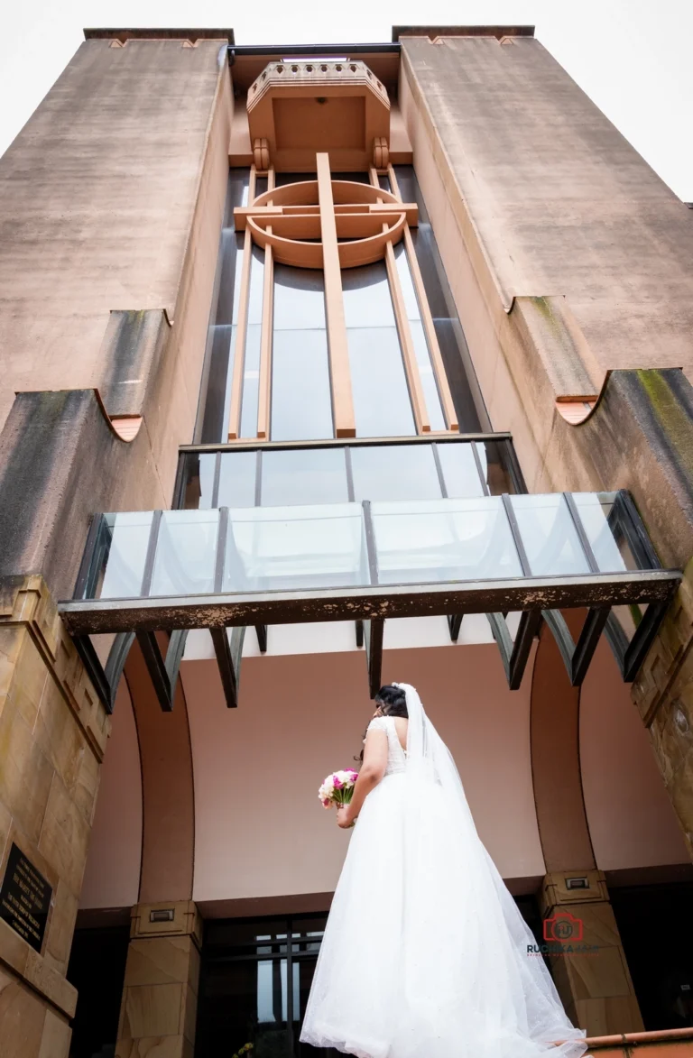 A bride in a white wedding gown holding a pink bouquet, standing beneath the grand architecture of the Wellington Cathedral of St. Paul