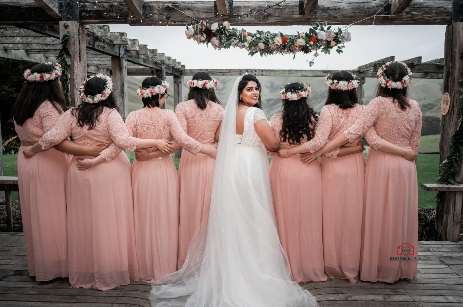 Bride in white gown stands with her back to the camera, arm-in-arm with her bridesmaids dressed in matching pink dresses and floral crowns, under a decorated pergola