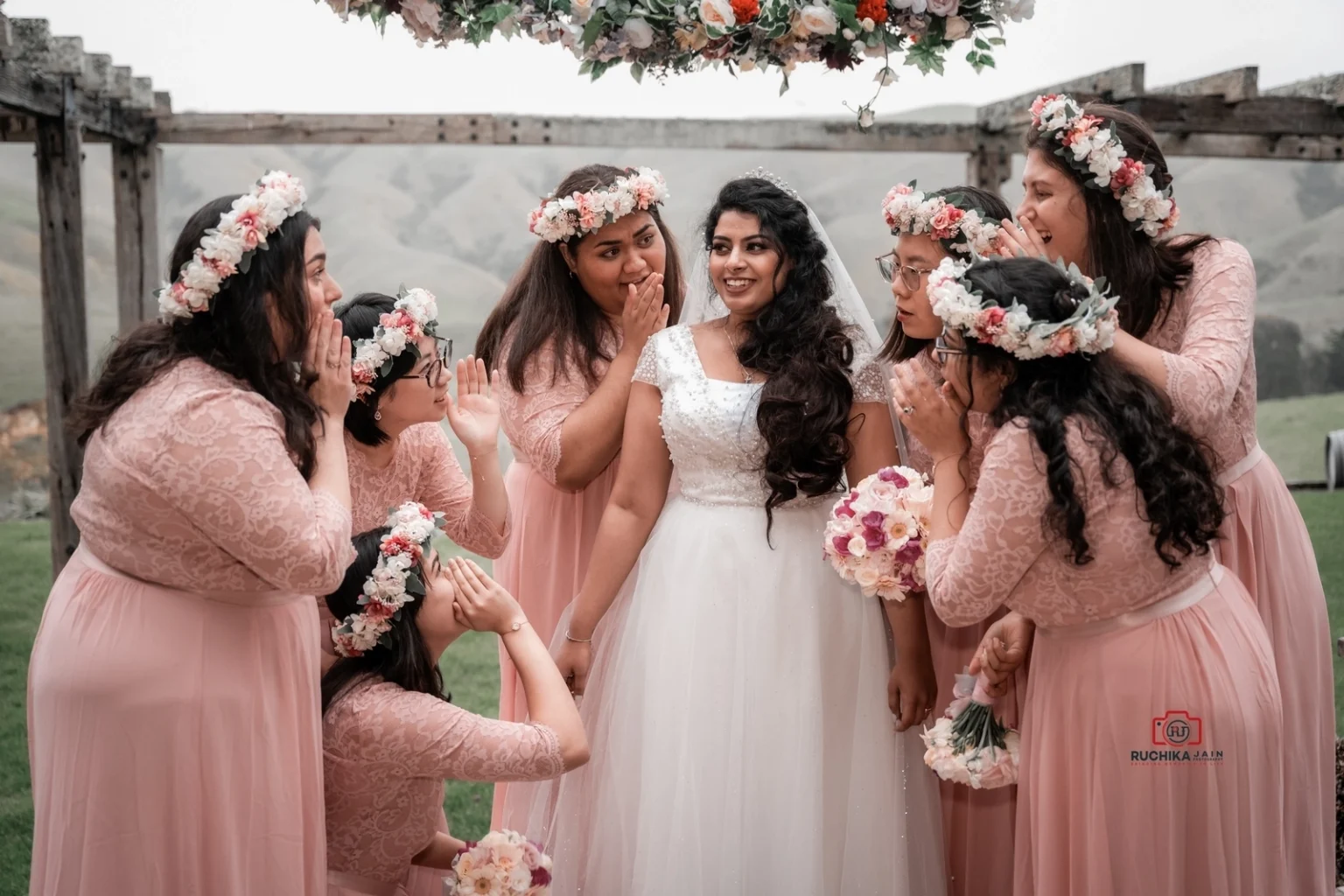 Bride in a white gown surrounded by bridesmaids in pink dresses, all looking playfully surprised with hands near their faces under a floral-decorated pergola