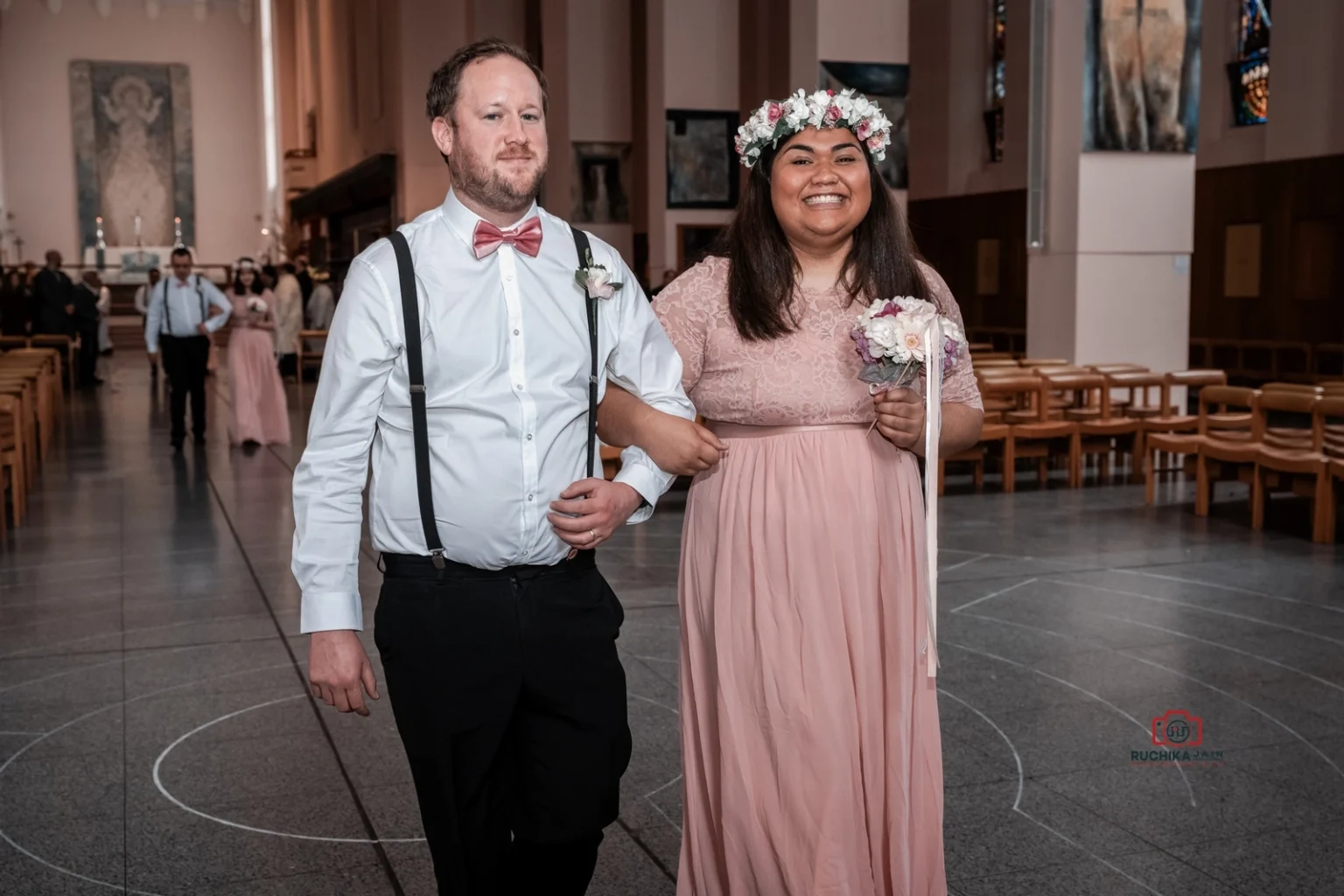 Bridesmaid in pink lace dress and floral crown smiles while walking arm-in-arm with groomsman in white shirt and suspenders